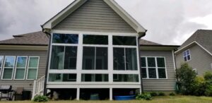 A serene gray house stands elevated, its bright sunroom with large windows overlooking lush greenery below.