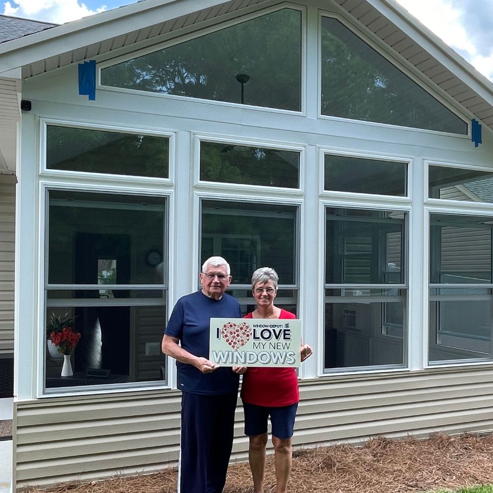 Smiling homeowners proudly hold an "I love my new windows" sign outside their freshly updated house.