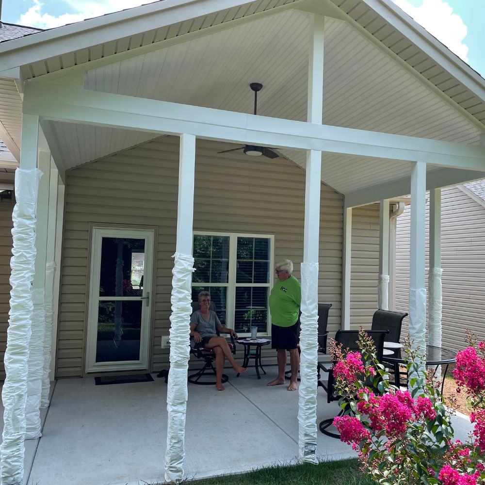 Two people converse on a shaded patio, surrounded by white posts and vibrant pink blooms.