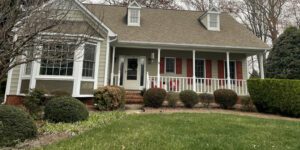 A charming two-story house with bay windows and a welcoming porch sits amid manicured bushes.