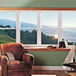 A cozy leather armchair sits by bay windows overlooking tranquil hills, trees, and sparkling water.