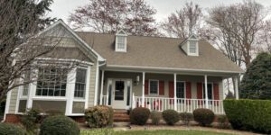 A charming beige house with red shutters and a welcoming porch sits amid neatly manicured bushes.