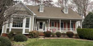 A cozy beige house with a welcoming porch sits amid a beautifully maintained, tree-lined yard.