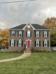 A charming brick house stands amid autumn trees, its red door inviting against a colorful seasonal backdrop.