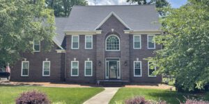 A stately brick home with steep roof overlooks a lush lawn, nestled among vibrant green trees.