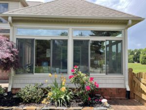 A bright sunroom with wide windows overlooks a peaceful garden bordered by greenery and a wooden fence.